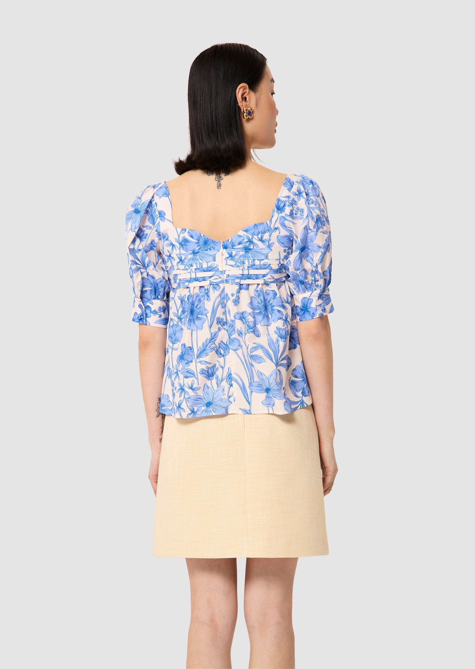 back view of a Woman wearing a blue floral top and beige skirt on a light gray background